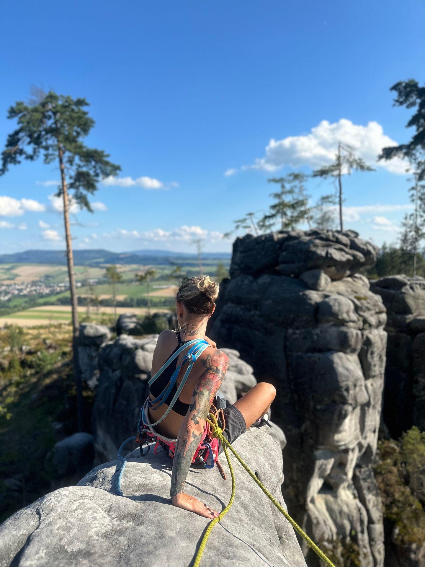 Person with tattoos sitting on a rocky cliff, secured with climbing gear, overlooking a scenic landscape.