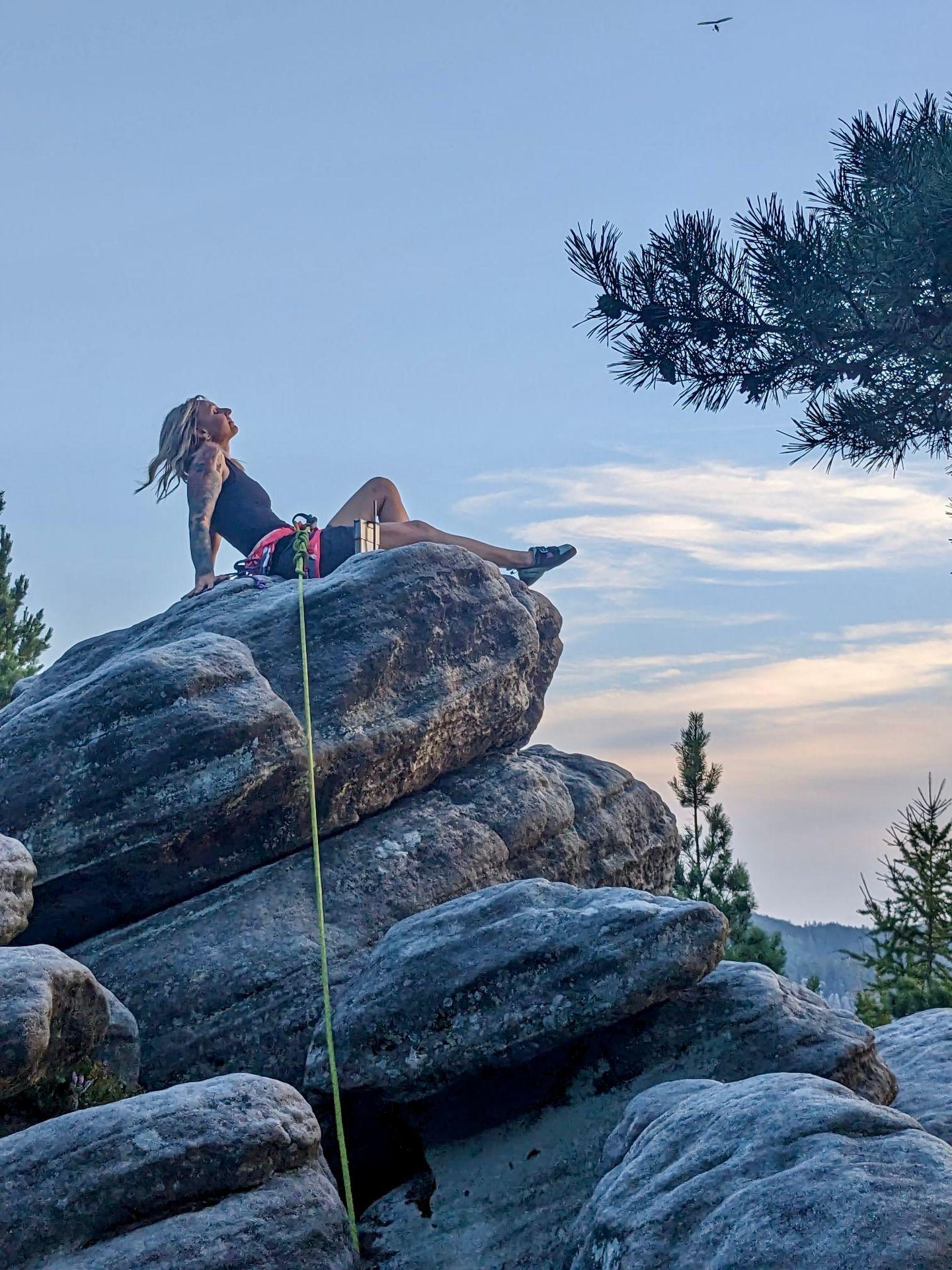 Person sitting on large rock under blue sky, surrounded by trees, with climbing gear visible.